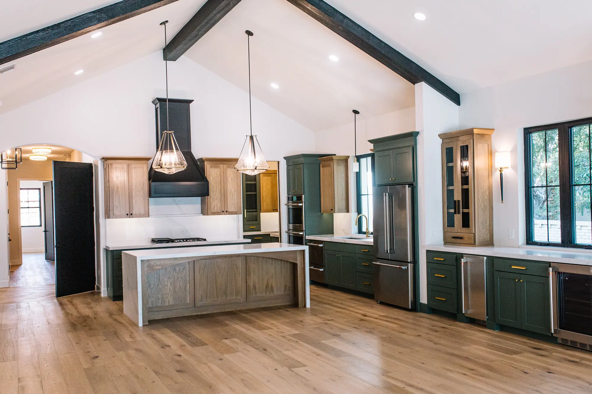 Open kitchen and living space under a cathedral ceiling with dark beams, with sage green cabinetry, a white oak island and engineered white oak flooring