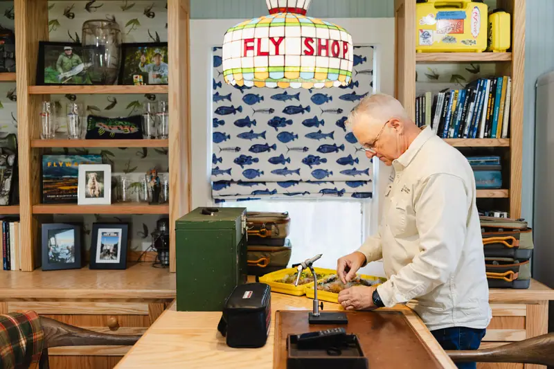 The homeowner tying flies at the furniture-style peninsula, with the custom built-ins, fish-inspired Roman shade and stained-glass Fly Shop chandelier behind him