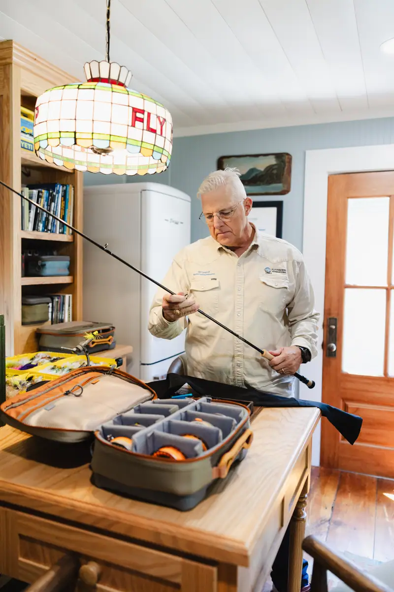 The homeowner checking a fly rod beneath the stained-glass Fly Shop chandelier, beside the vintage refrigerator and the wood entry door