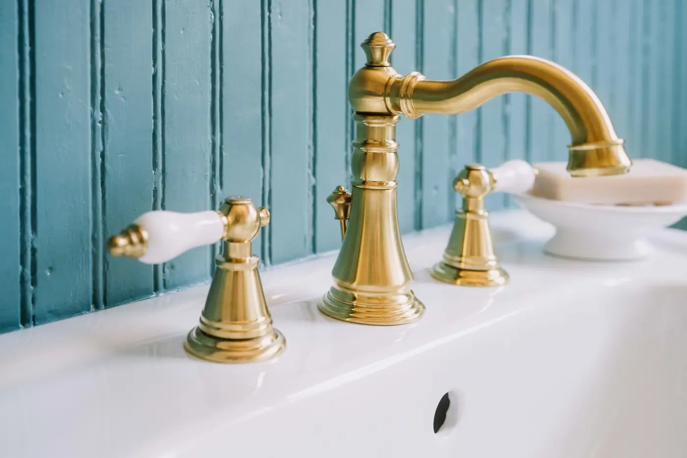 A brass widespread faucet with porcelain levers on a white sink, against teal beadboard panelling.