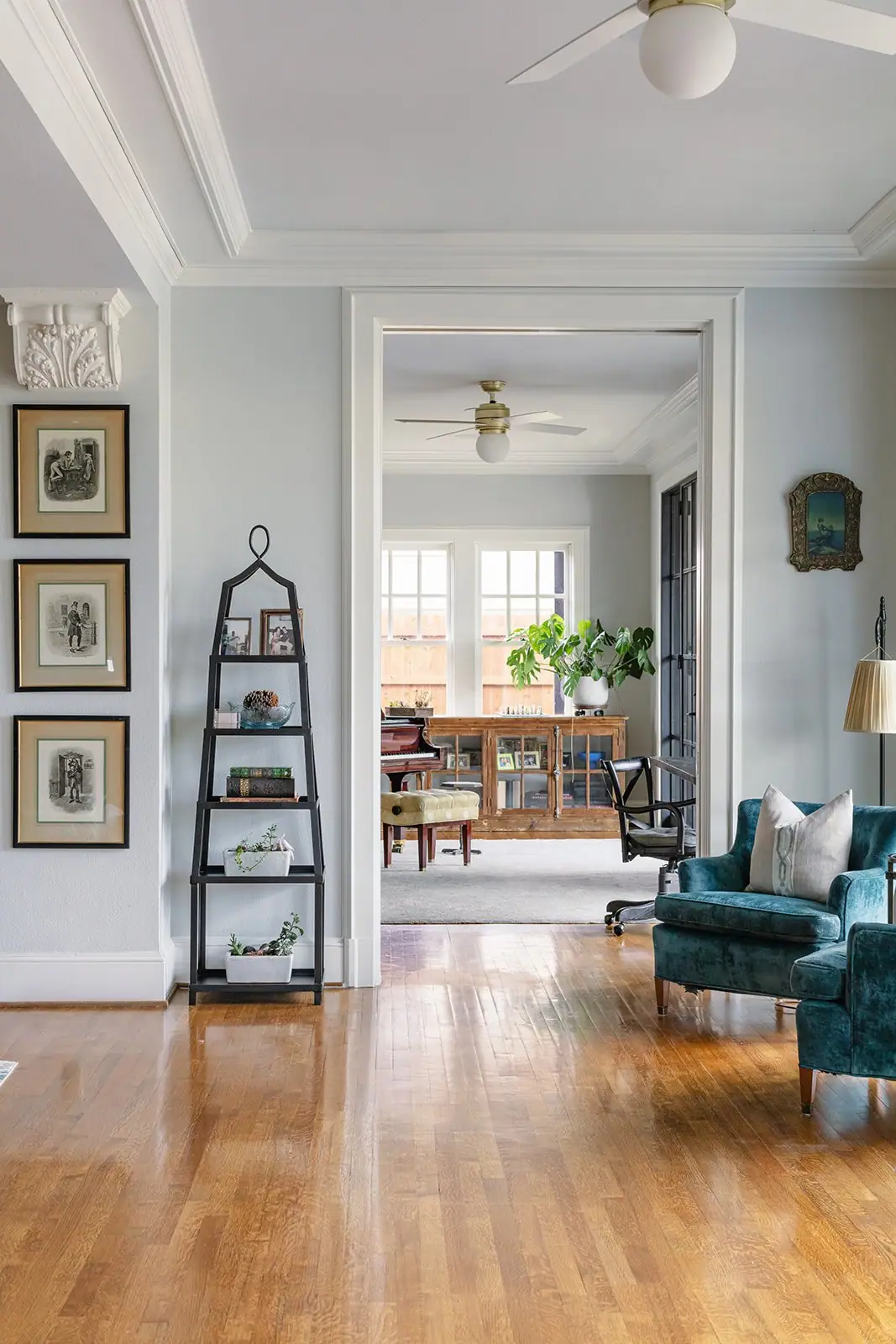 Living room with teal velvet chairs and framed prints, looking through a wide cased opening to a room with a grand piano