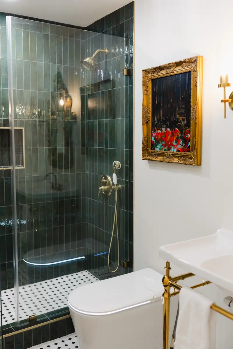Bathroom with a deep green glazed tile shower, a patterned mosaic floor, brass fixtures and a gilt-framed painting