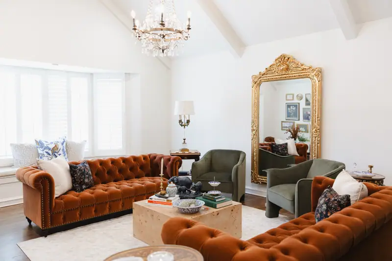 Living room looking toward a bay window, with two rust velvet chesterfield sofas, green velvet chairs and a tall gilt leaning mirror