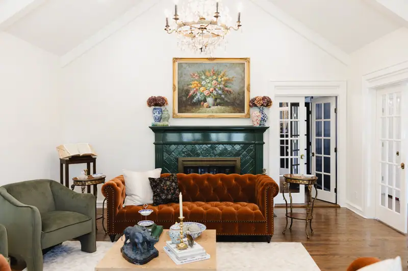 Living room with a vaulted ceiling, crystal chandelier, deep green tiled fireplace, rust velvet chesterfield sofa and French doors