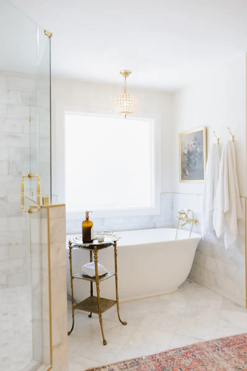 Primary bathroom wrapped floor to ceiling in Carrara marble, with a freestanding tub, brass fixtures and a glass shower