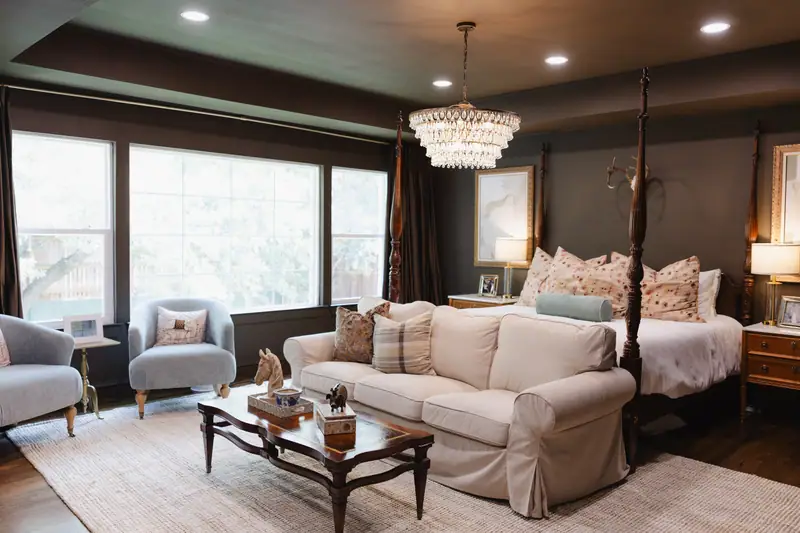 Primary bedroom drenched in chocolate brown across the walls and ceiling, with a four-poster bed, crystal chandelier and a sitting area by the windows