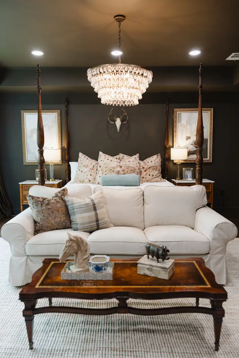 Primary bedroom looking toward the four-poster bed, with chocolate brown walls, a tiered crystal chandelier and framed art flanking the bed