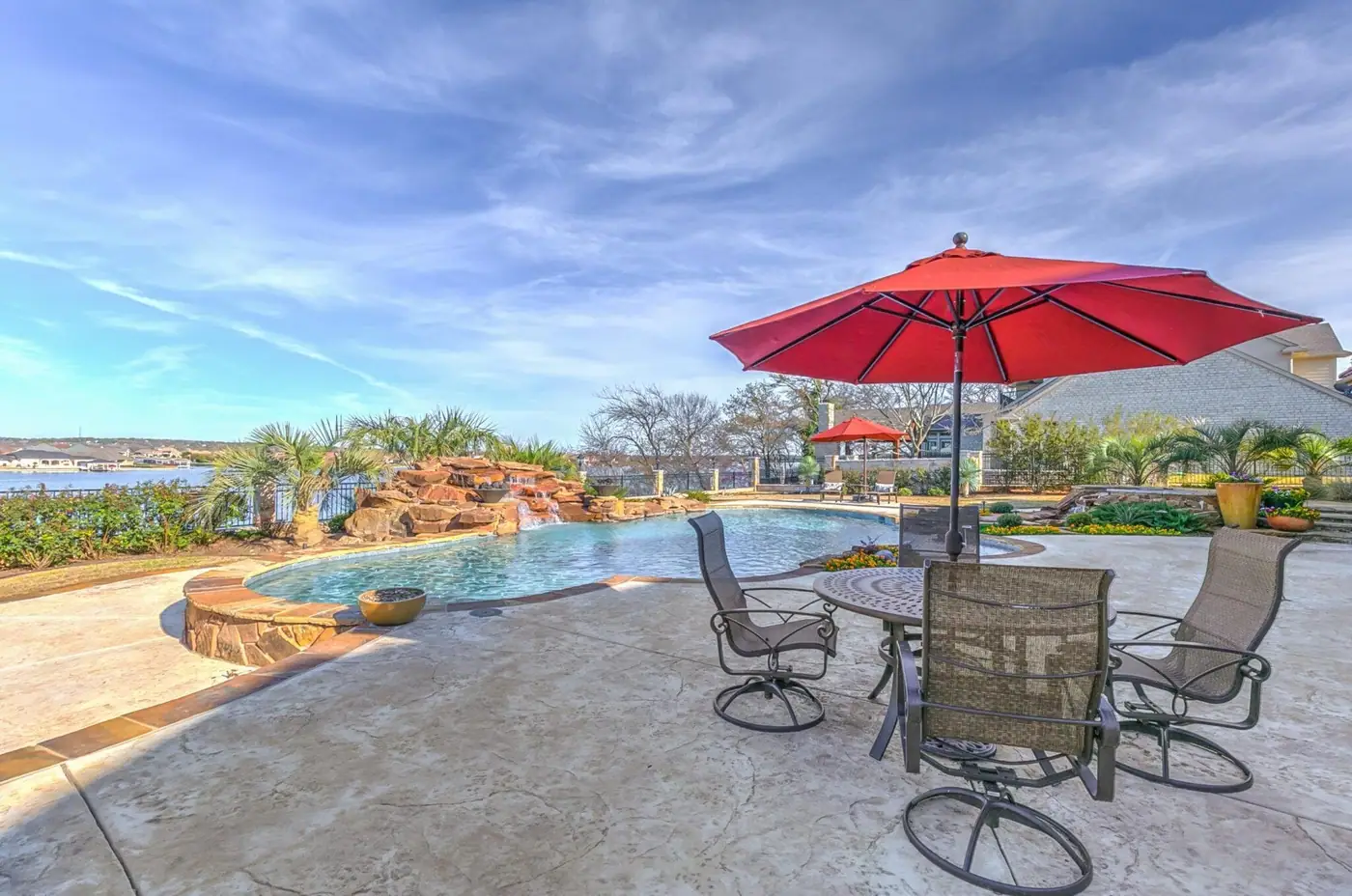 A free-form pool with a boulder waterfall at its far end, seen across a stamped deck from under a red umbrella