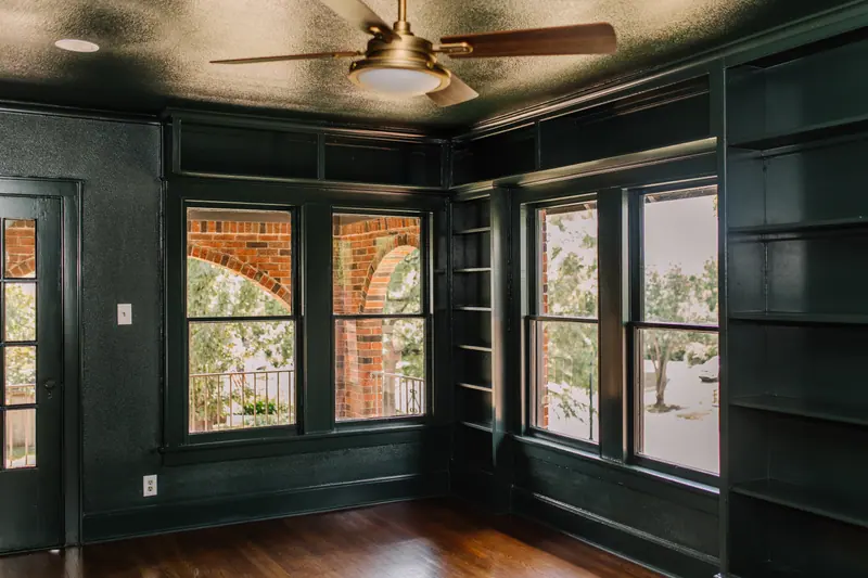 A room painted deep green from the floor to the ceiling, built-in shelves and window trim included, with a brass ceiling fan and two pairs of windows onto the arched brick porch
