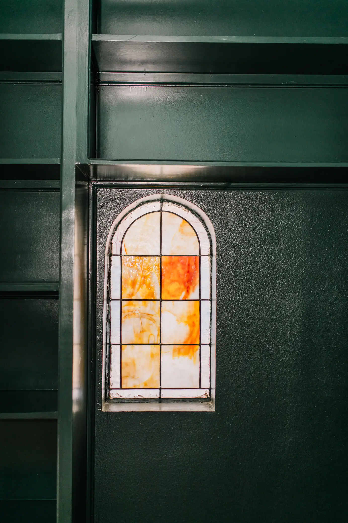 An arched stained-glass window in amber and red, set into a wall painted the same deep green as the trim and built-in shelving around it