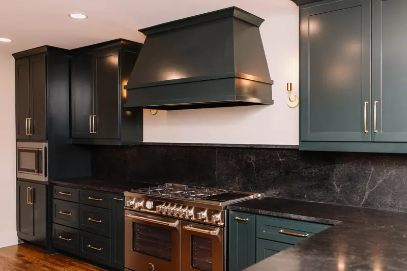 The kitchen: deep forest-green cabinets with brass pulls, a green range hood over a stainless range, and dark honed-stone counters and backsplash