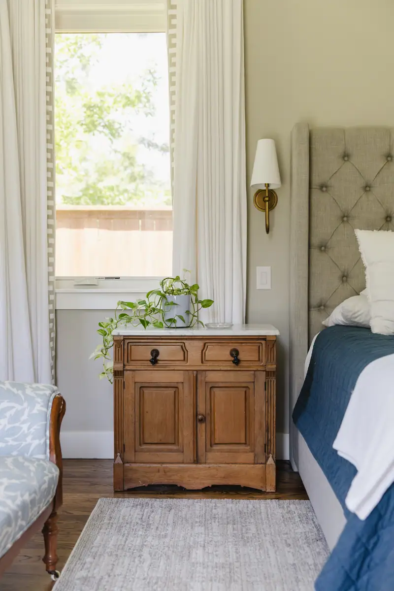 An antique washstand with a marble top as a nightstand, under a brass sconce beside the tufted headboard and the bordered drapery