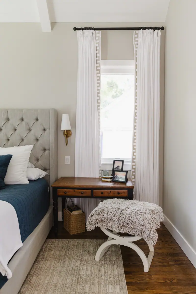 A bedside corner in the primary bedroom: a tufted linen headboard, a brass sconce, a wood writing desk as a nightstand and Greek-key drapery at the window