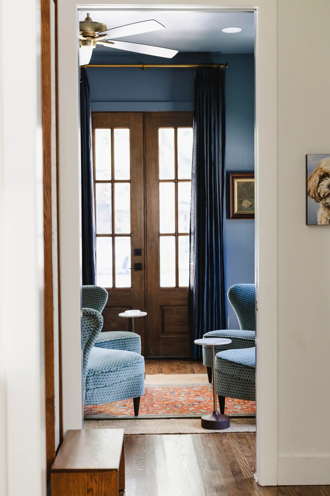 The smoky blue den of a Tanglewood custom home in Fort Worth, seen through its doorway, with navy drapery, blue velvet chairs and wood-and-glass doors