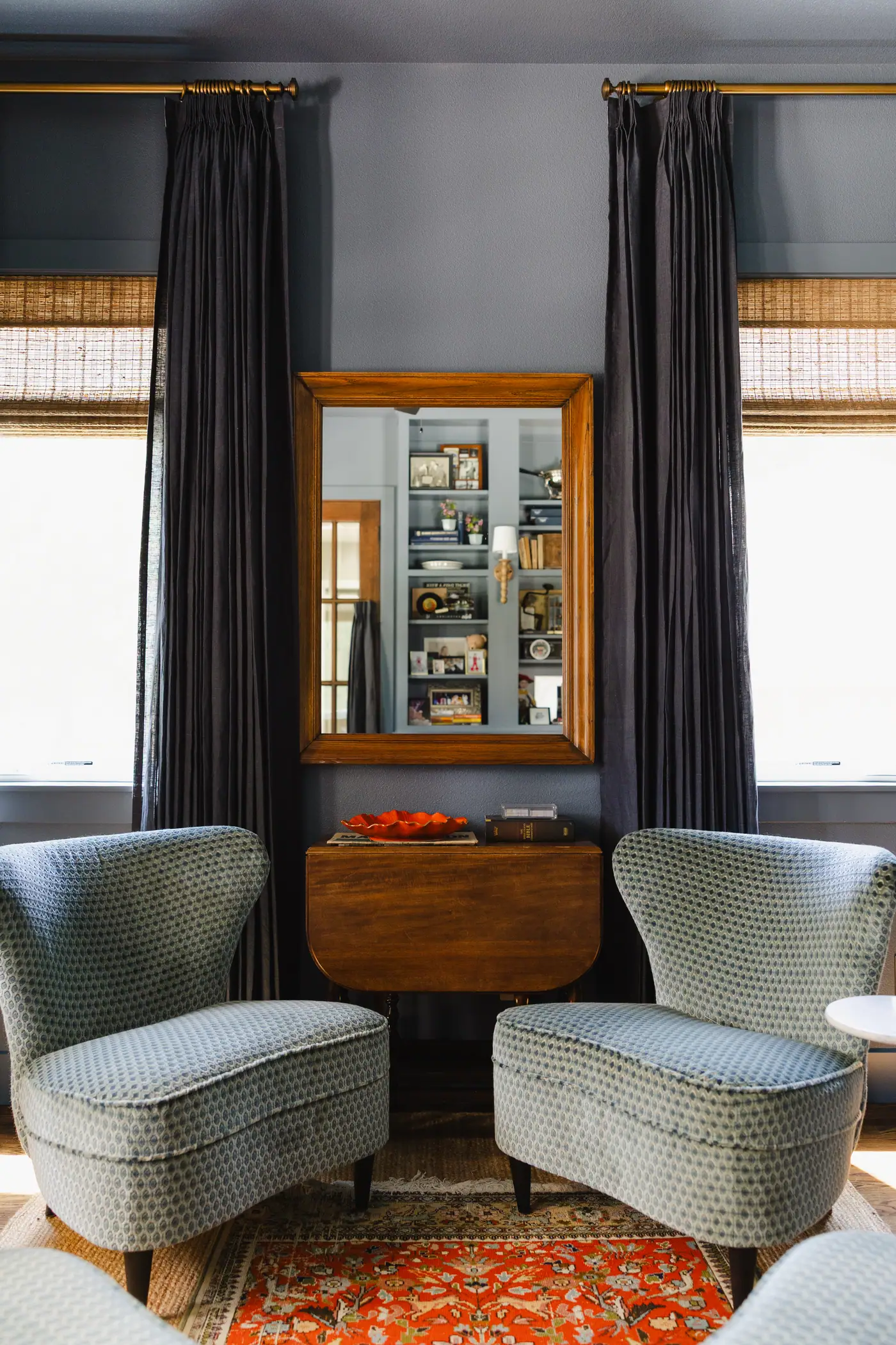 The den's window wall: navy drapery on brass rods over woven shades, a gilt mirror above a drop-leaf table, and a pair of patterned blue lounge chairs on a red rug, the walls and ceiling in one smoky blue