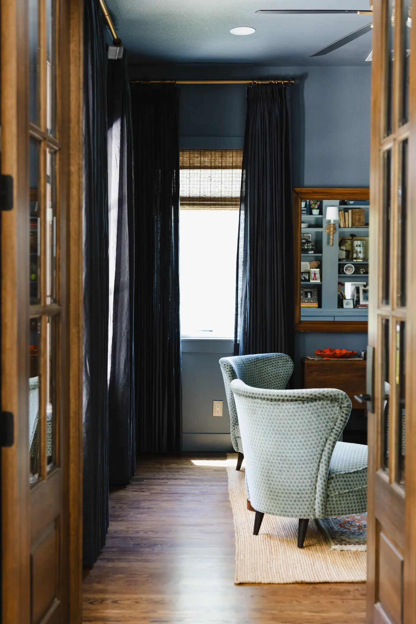 Smoky blue walls and ceiling, navy drapery over a woven shade, a pair of patterned blue lounge chairs and a built-in shelf wall