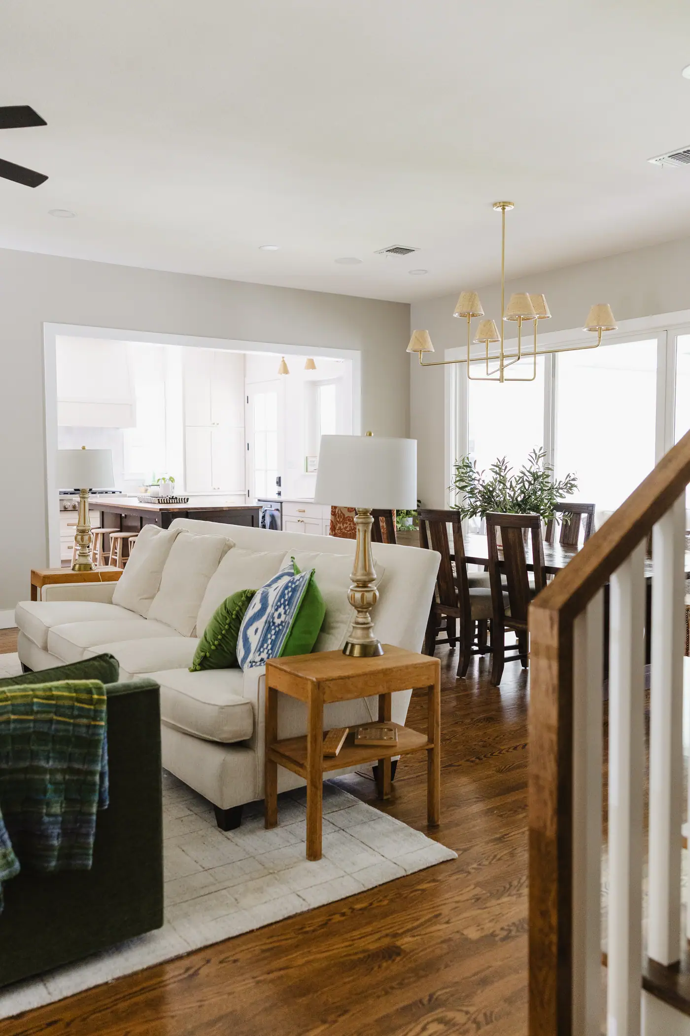 The living room open to the dining room: a cream sofa with blue and green pillows, a green velvet chair, a wood side table and brass lamp, and a brass chandelier over the dining table beyond