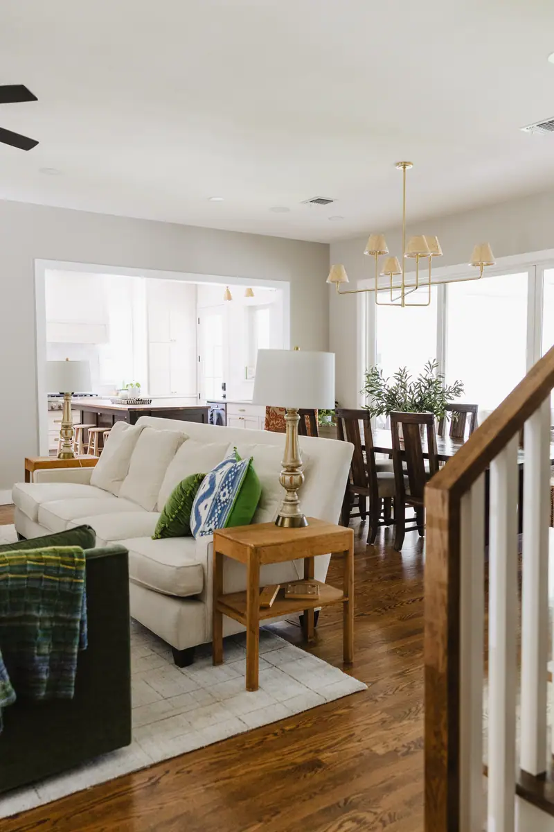 The living room open to the dining room: a cream sofa with blue and green pillows, a green velvet chair, a wood side table and brass lamp, and a brass chandelier over the dining table beyond