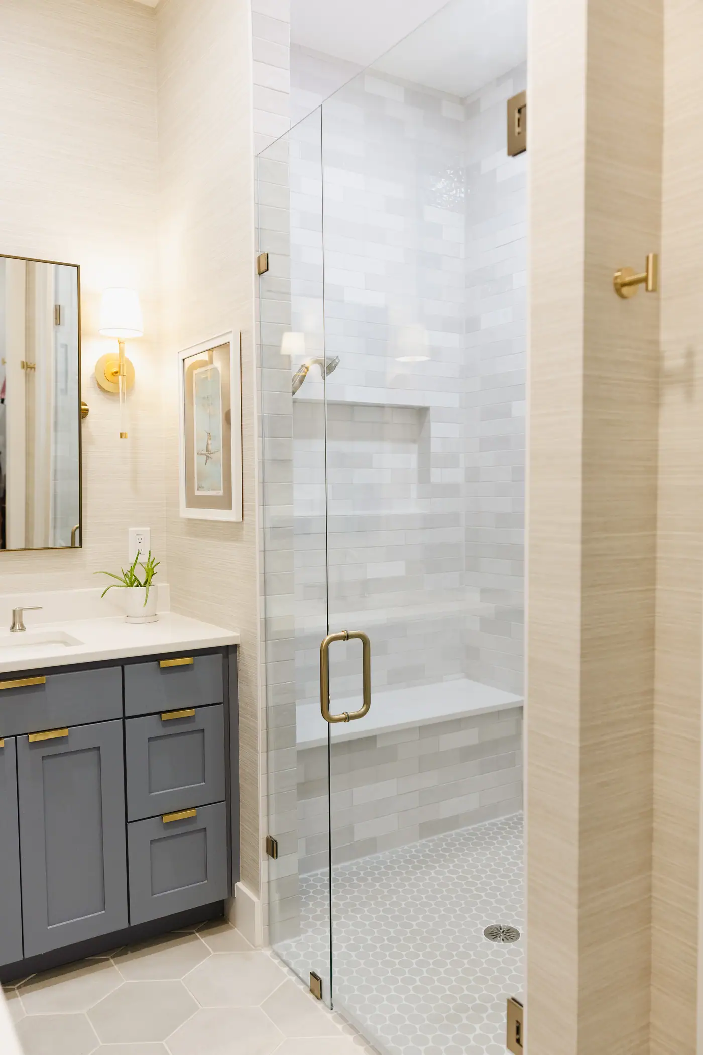 The primary bath shower, glass-enclosed in pale stacked tile with a bench and niche, beside the smoky blue vanity and grasscloth walls