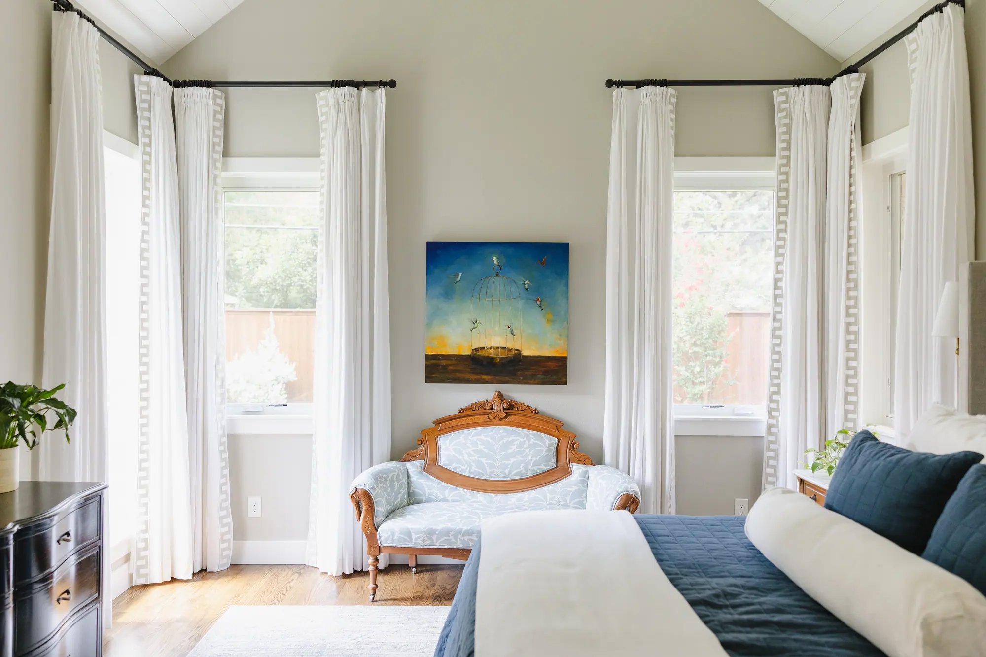 The primary bedroom: tall white drapery with a Greek-key border hung high on black rods, an antique carved settee under a painting of hummingbirds, and a navy quilt on the bed