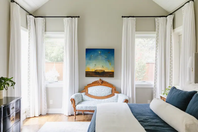 The primary bedroom: tall white drapery with a Greek-key border hung high on black rods, an antique carved settee under a painting of hummingbirds, and a navy quilt on the bed