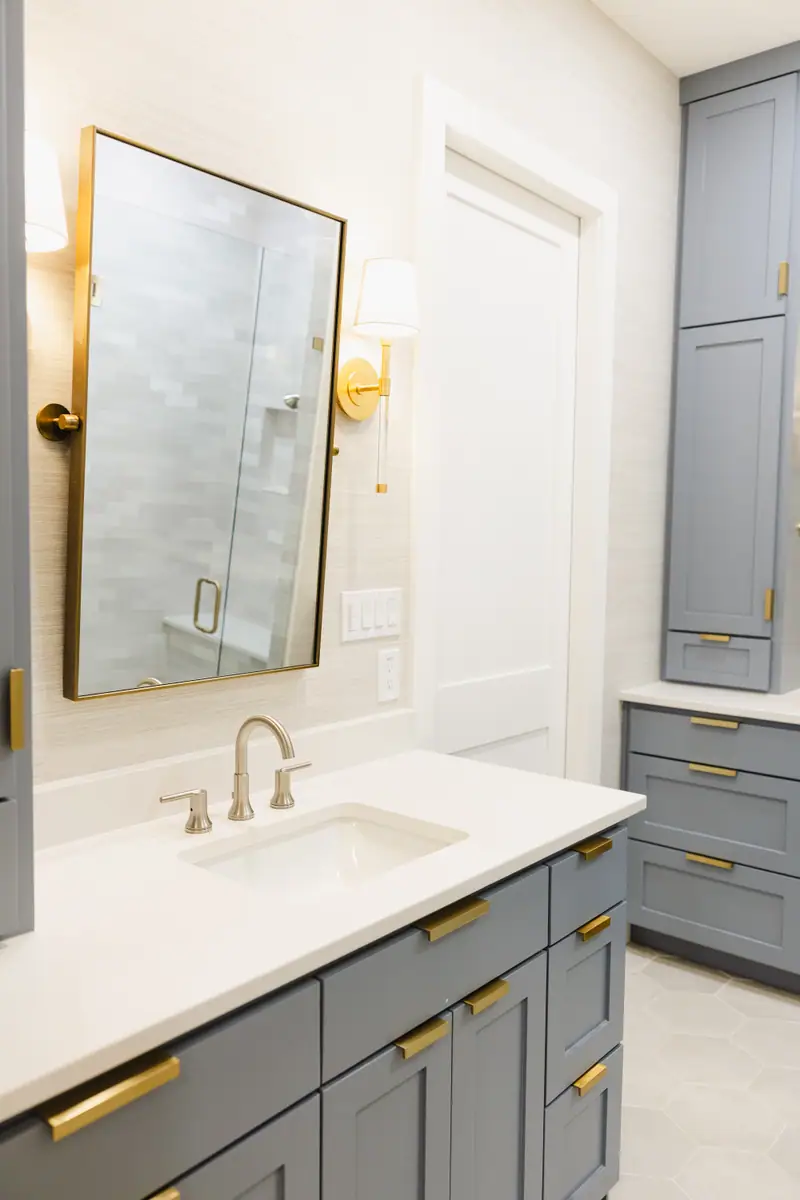 A bathroom vanity in smoky blue cabinetry with brass pulls, a brass-framed mirror and brass sconces against textured wallcovering