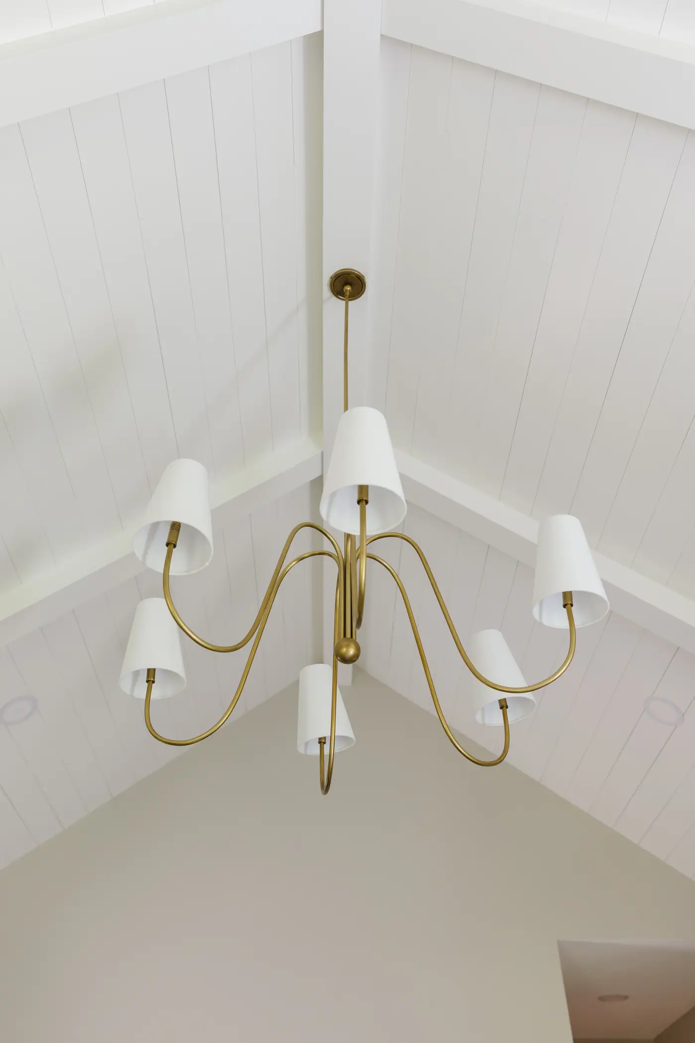 A brass chandelier with white shades hung at the peak of the primary bedroom's white shiplap vaulted ceiling