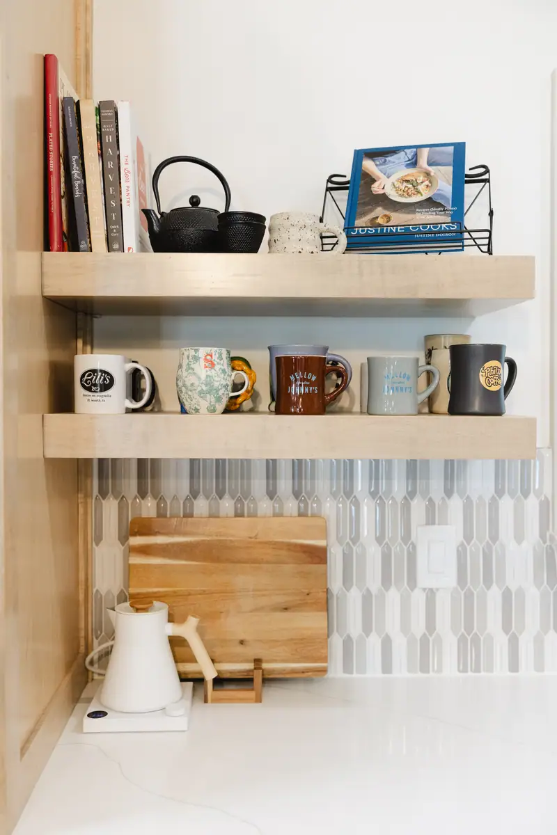 Floating light wood shelves over a picket tile backsplash, styled with cookbooks and mugs