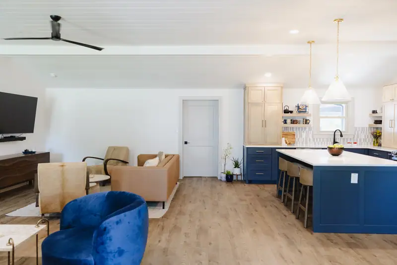 Living area with a tan leather sofa and a blue velvet chair, opening to the kitchen and its navy island beyond