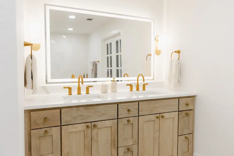 Primary bathroom double vanity in light wood with brass fixtures, wall sconces and a backlit mirror