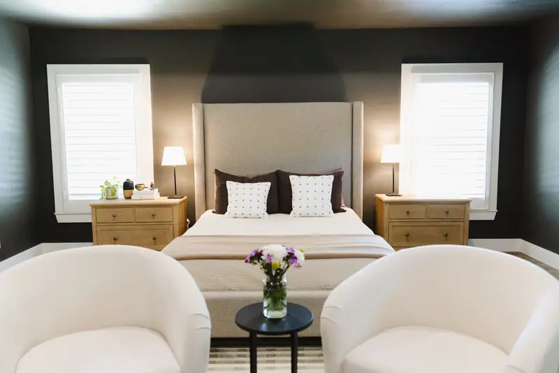 Primary bedroom bed with a tall upholstered headboard between two wood nightstands and lamps, against a dark wall