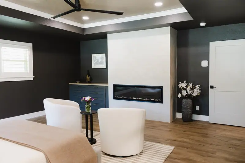 Primary bedroom with dark walls, a linear fireplace, a navy built-in dresser and a pair of ivory swivel chairs