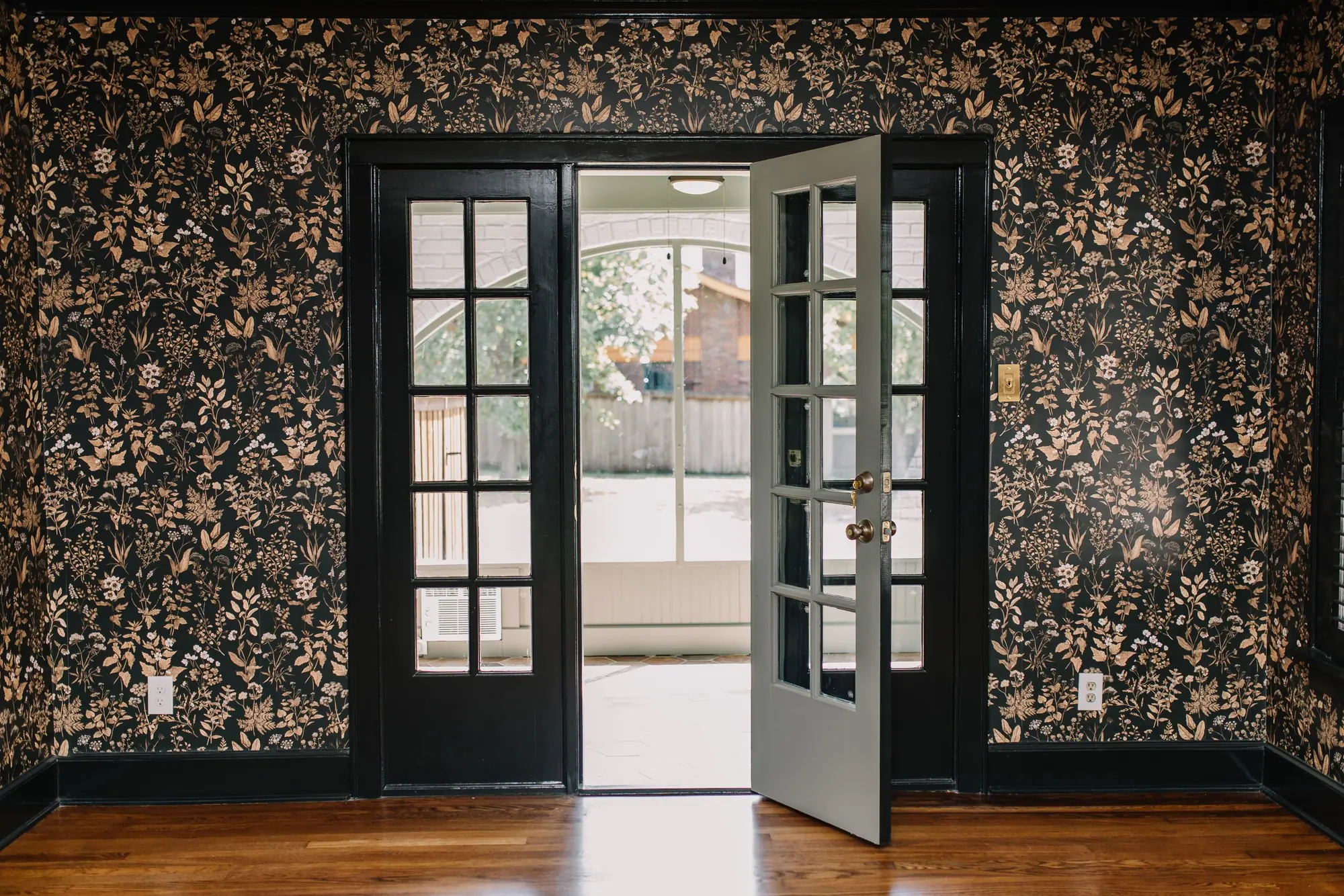 A Fort Worth whole-home remodel: black French doors standing open off a room papered in dark botanical print, with oak floors running through